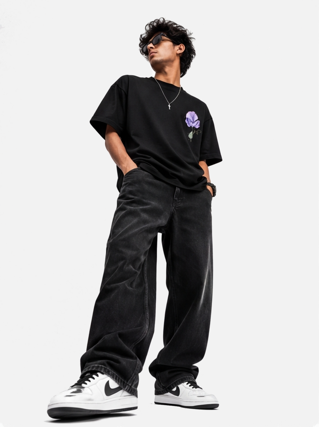 A model wearing a black oversized t-shirt with a purple rose graphic on the chest, paired with black wide-leg jeans and white sneakers, standing against a light gray background.